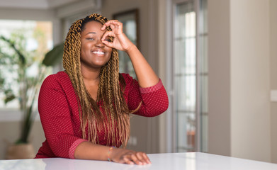 Wall Mural - African american woman sitting at home with happy face smiling doing ok sign with hand on eye looking through fingers