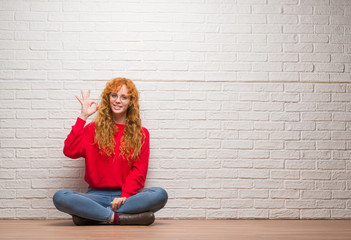 Poster - Young redhead woman sitting over brick wall smiling positive doing ok sign with hand and fingers. Successful expression.