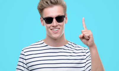 Wall Mural - Young handsome blond man wearing sunglasess surprised with an idea or question pointing finger with happy face, number one