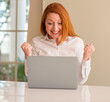 © Krakenimages.com - Redhead woman using computer laptop at home screaming proud and celebrating victory and success very excited, cheering emotion