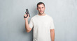 © Krakenimages.com - Young caucasian criminal man over grey grunge wall holding gun with a confident expression on smart face thinking serious