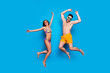 Full length portrait of a joyful young jumpers woman and happy man in sunglasses, dressed in swimsuit, jumping and putting hands up over blue background. Beach party style!