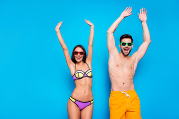 Poster - Hot summer time concept. Portrait of cheerful couple in sunglasses and bright swimsuits enjoying sunlight together with beach party mood raise their hands up isolated on bright blue background