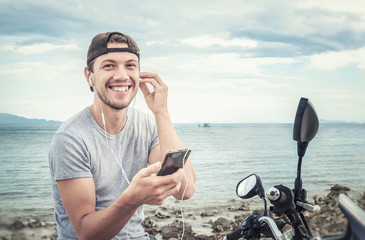 Young smiling man with smartphone and earphones on the sea coast