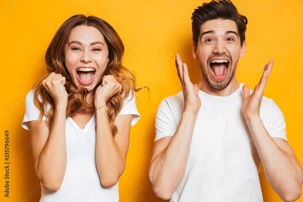 Photo of beautiful admired couple man and woman in basic clothing screaming in surprise or delight and touching cheeks, isolated over yellow background - obrazy, fototapety, plakaty 