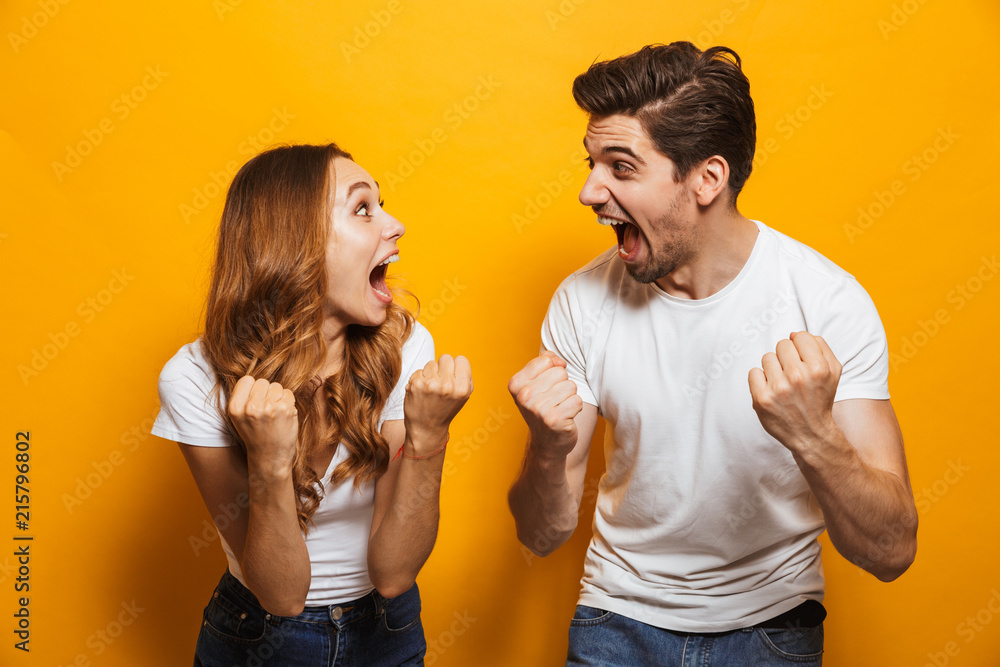 Photo of delighted european man and woman in basic clothing screaming and clenching fists like winners or happy people, isolated over yellow background - obrazy, fototapety, plakaty 