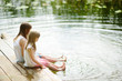 © MNStudio - Two cute little girls sitting on a wooden platform by the river or lake dipping their feet in the water on warm summer day