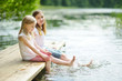 © MNStudio - Two cute little girls sitting on a wooden platform by the river or lake dipping their feet in the water on warm summer day