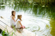 © MNStudio - Two cute little girls sitting on a wooden platform by the river or lake dipping their feet in the water on warm summer day