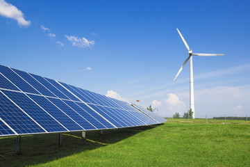 wind generator of electricity from three blades and solar panels of a battery of photocells against a background of clouds and a blue sky green grass