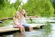 © MNStudio - Two cute little girls sitting on a wooden platform by the river or lake dipping their feet in the water on warm summer day