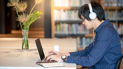 Wall Mural - Young Asian man working with laptop computer listening to music in public library. Male university student doing research in college building. Education and technology concepts