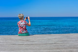 Fototapeta Pomosty - Kobieta z aparatem fotograficznym, siedząca na drewnianym pomoście nad morzem