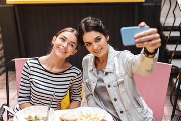 Wall Mural - Selfie together. Two beaming appealing women wearing stylish clothes making selfie together in cafeteria