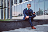 Fototapeta Pomosty - Portrait of a confident stylish businessman dressed in an elegant suit holds a smartphone and looking at camera while sitting outdoors against a skyscraper background.