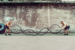 © zamuruev - Two muscular athletes training, working out with rope in front of brick wall. Scream. Working hard. Street gym. Strength and motivation. Outdoor workout.