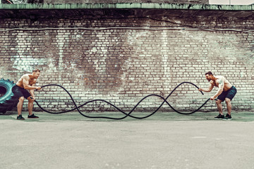 Two muscular athletes training, working out with rope in front of brick wall. Scream. Working hard. Street gym. Strength and motivation. Outdoor workout.