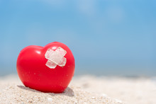 Stick Plaster On Broken Heart. Plaster On Red Heart On Sand With Blue Sky Background. Heart Repaired And Recovery.
