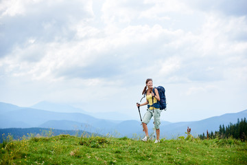 Sporty smiling woman backpacker hiking in Carpathian mountain trail, walking on grassy hill, wearing backpack, using trekking sticks, enjoying summer cloudy day. Outdoor activity, lifestyle concept