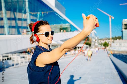 Fotografía Young hipster woman traveler walking in Oslo city on quay, listening to music with red headphones, street style, urban Oslo Opera House background, holding smart phone, taking selfie photo