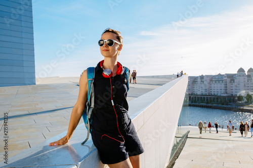 Photo Young hipster woman traveler in sunglasses walking in Oslo city summer sunny day with red headphones, backpack, street style, Opera House urban background, , happy, smiling