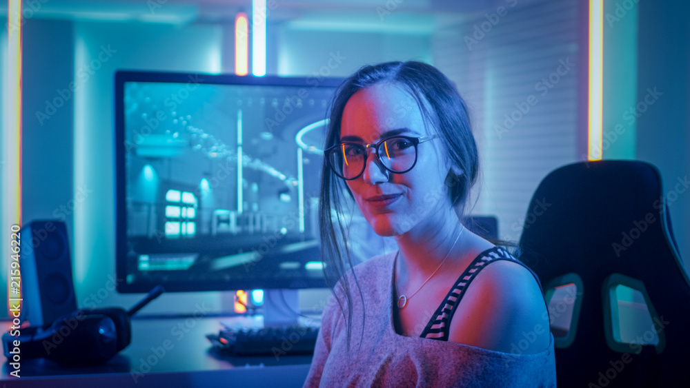 Portrait of the Beautiful Young Pro Gamer Girl Sitting at Her Personal Computer and Looks into Camera. Attractive Geek Girl Player Wearing Glasses in the Room Lit by Neon Lights. - obrazy, fototapety, plakaty 