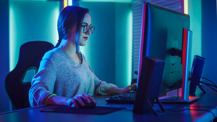 Beautiful Professional Gamer Girl Sitting Down to Play in First-Person Shooter Online Video Game on Her Personal Computer. Casual Cute Geek wearing Glasses and Talking into Headset. Cyber e-Sport.