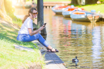 Outdoor summer smiling lifestyle portrait (horizontal) of young pretty brunette woman traveling in Giethoorn (Netherlands) and making picture (with smartphone)of floating in the channel duck.