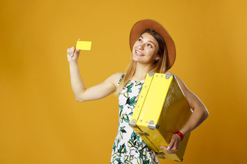 Young happy woman holding empty credit card in one hand and yellow suitcase in another over orange background