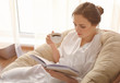 © Pixel-Shot - Young woman drinking coffee while reading book on lounge chair at home