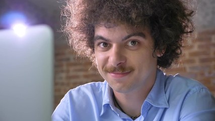 Funny nerd hacker typing on laptop and looking at camera smiling, brick wall background