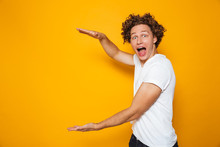 Young Excited Man 20s With Brown Curly Hair Gesturing Aside And Holding Big Copyspace In Hands, Isolated Over Yellow Background