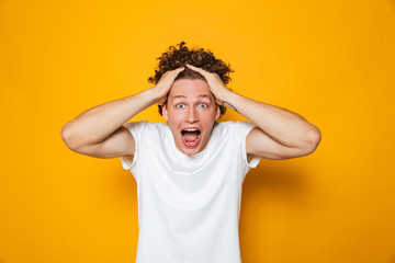 Poster - Portrait of emotional shocked guy with curly brown hair screaming and grabbing head, isolated over yellow background