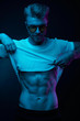 © monchak - Neon light portrait of sexy man in yellow glasses, pulling up t-shirt, showing abs