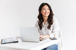 © Drobot Dean - Photo of charming chinese businesswoman with long dark hair sitting at table and working with documents and laptop, isolated over white background in studio