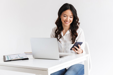 Poster - Photo of young asian businesswoman with long dark hair sitting at table and holding smartphone while working with laptop, isolated over white background in studio