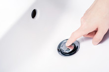 Female Hand With Finger On An Open, Pop-Up Drain Plug, Ready To Press And Close The Drain