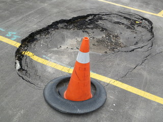 Constructions at unexpected sink hole in public parking lot with traffic cone. This accident causes damage or injuries. It needs instant attention and repair work and proper foundation of the ground.