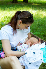 Detail of female parent nursing the newborn, young happy mother is breastfeeding the infant, she is holding the minor gently in the hands with uncovered breast, outside city park scene