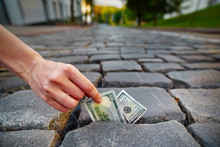Hand Taking A Hundred-dollar Bill, Lying On The Pavement. The Concept Of Corruption And Theft In The Construction And Maintenance Of Roads. Investments In The Construction And Maintenance Of Roads.
