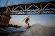 © fesenko - Young man jumping on wakesurf down the river waves against the bridge at the sunset