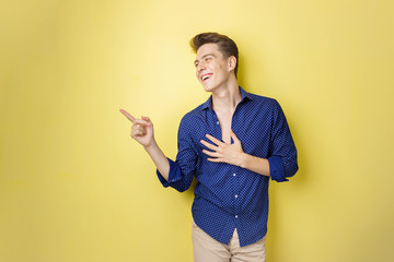 Friendly looking cheerful european guy with dark hair wearing blue shirt, smiling widely, pointing left while standing over yellow background