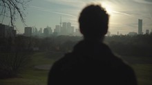 Young Adult Man Stands And Looks At City Skyline From A Distance, Silhouette Of Millennial In A Park On Beautiful Sunny Evening At Golden Hour Before Sunset, Cinematic Slow Motion