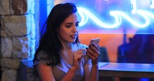Girl Using Cellphone At Night Next To Blue Neon Sign Messaging And Reading Texts