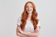 Portrait of cheerful pretty redhead young woman with long wavy hair and freckles wears t shirt keeps hands folded and laughing isolated over white background
