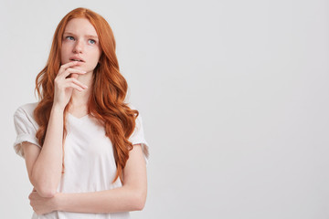 Portrait of pensive attractive redhead young woman with long wavy hair and freckles wears t shirt touching her chin and thinking about future isolated over white background