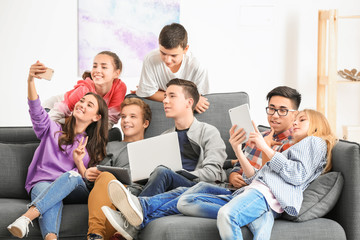 Wall Mural - Group of teenagers with modern devices taking selfie while sitting on sofa indoors