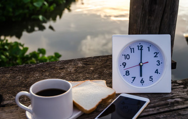 Coffee bread and White clock on a wooden table in the morning.On the river at 6:00 pm, there is a white clock on the wooden floor.Smartphone put on table.Do not focus on objects.Warm tone.