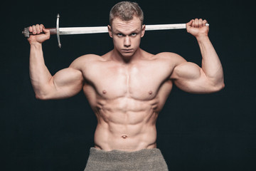 Wall Mural - Bodybuilder man posing with a sword isolated on black background. Serious shirtless man demonstrating his mascular body