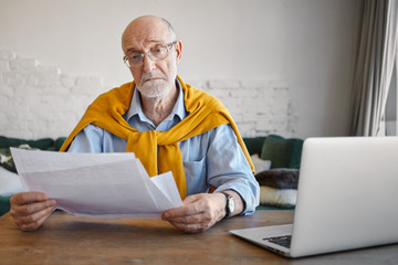 Wall Mural - Portrait of serious successful elderly male entrepreneur wearing stylish outfit and accessories checking financial papers in his hands, while working in modern office, using electronic device